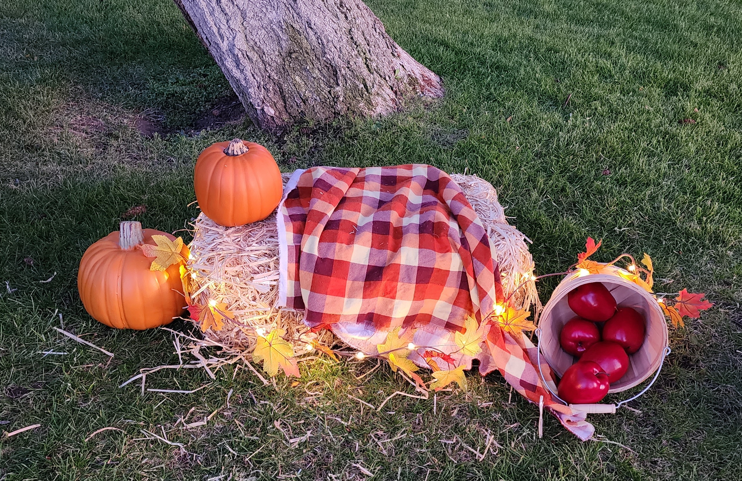 Straw bale covered in a fall flannel blanket and a string of leaves and LED lights with pumpkins and a bushel of apples on the ground beside it